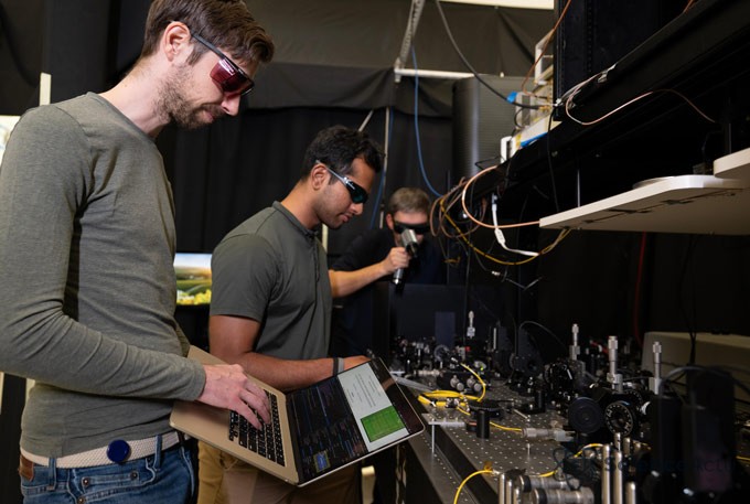 Trois hommes dans un laboratoire, l'un tenant un ordinateur portable, travaillant sur un protocole de génération de nombres aléatoires.