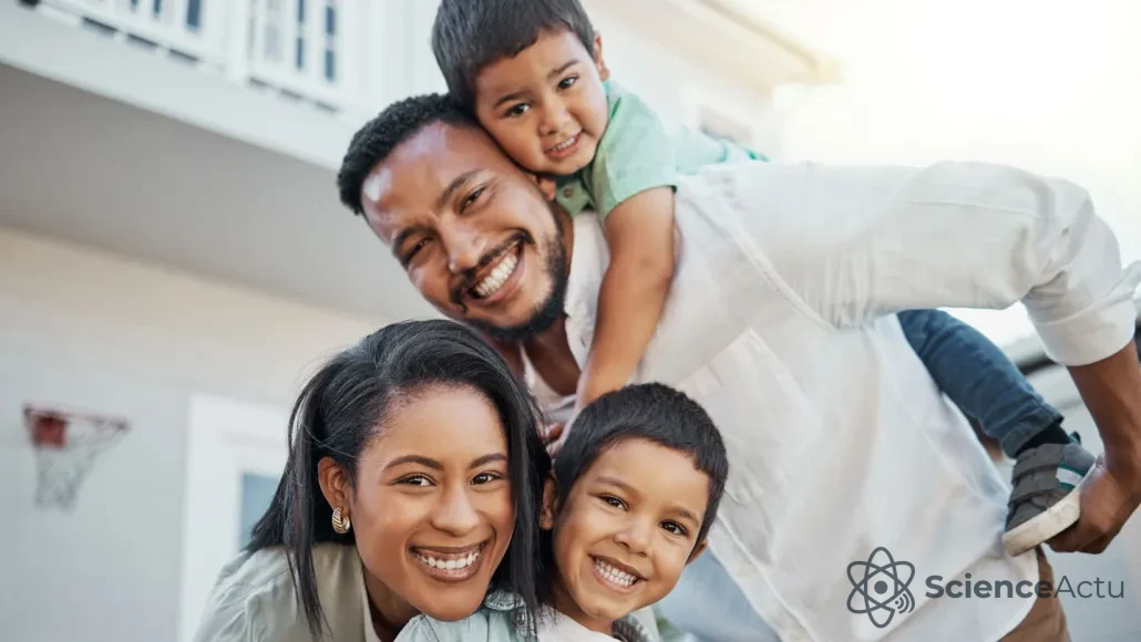 Famille souriante de quatre personnes à l'extérieur, avec un homme portant un jeune garçon sur son dos, tandis qu'une femme se penche à côté d'eux, tenant un autre garçon souriant devant elle.