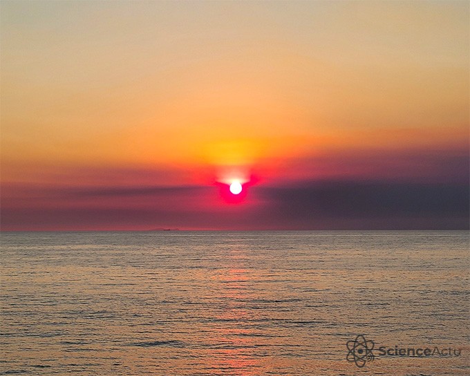 Le soleil légèrement au-dessus de la ligne de flottaison de l'océan. La fumée nuageuse rend le soleil couchant brumeux.