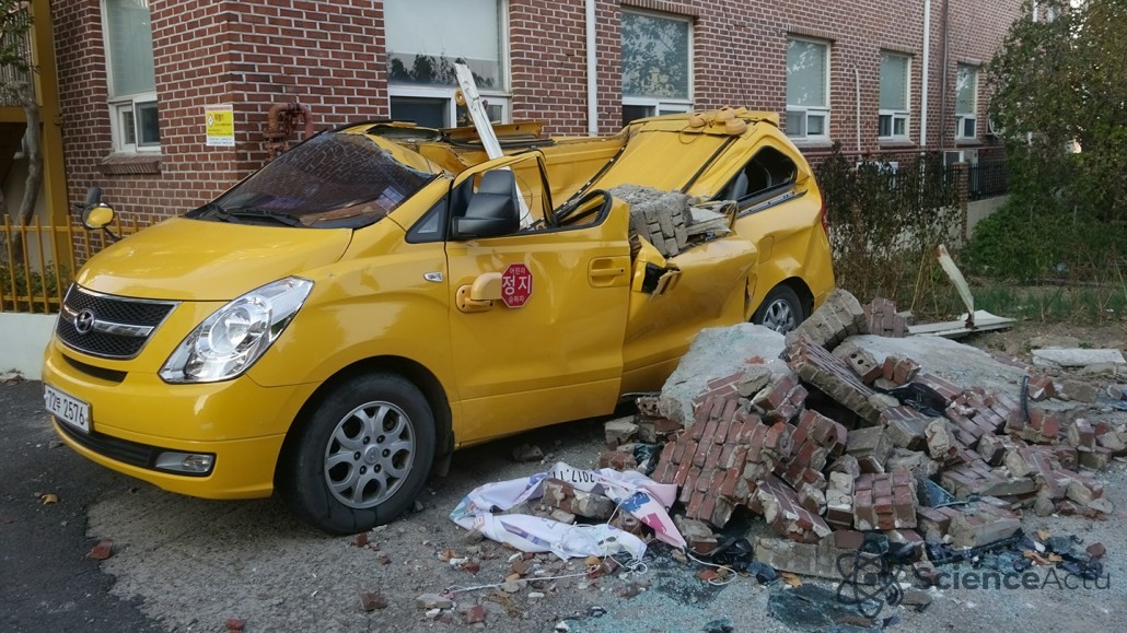 Un véhicule jaune écrasé par des briques et des débris devant un bâtiment en briques après un séisme.