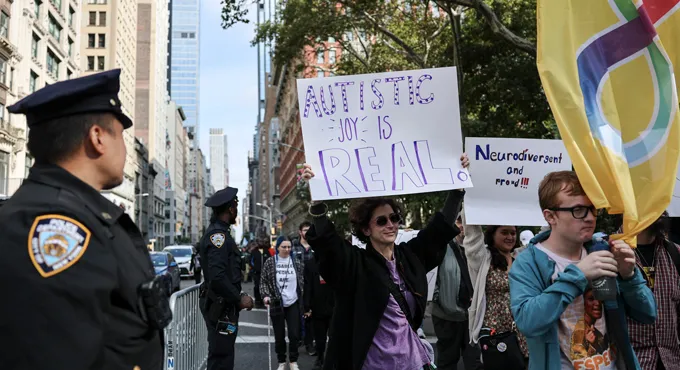 Une personne tient une pancarte indiquant 'Autistic joy is real' dans une rue de New York, face à un policier.